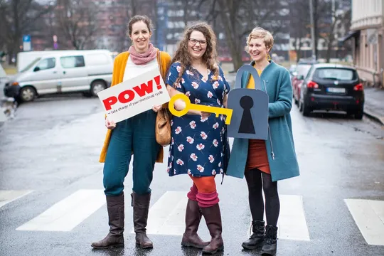 Queen Raae and two co-founders on a wet Oslo crosswalk, laughing and holding a POW! sign, a big yellow key and a cardboard padlock