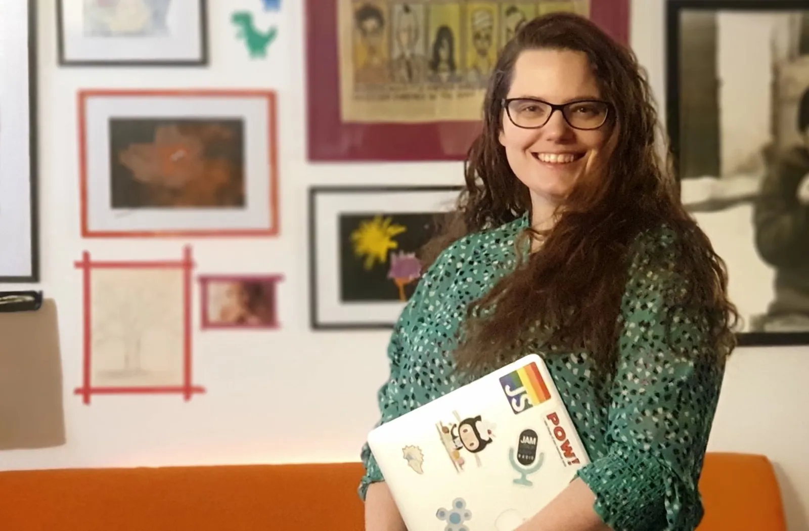 Queen Raae smiling in front of her gallery wall at home, holding a sticker-covered laptop