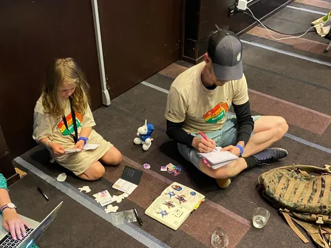 Ola and our daughter sitting on a conference hall floor drawing in notebooks, with Queen's laptop in the corner