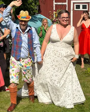Queen Raae in a lace wedding dress and Ola in flower shorts and a straw hat walking hand in hand across a lawn as guests cheer